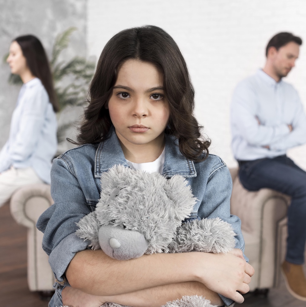 Girl holding a teddy bear with two adults seated apart behind her, illustrating child custody and parenting time.